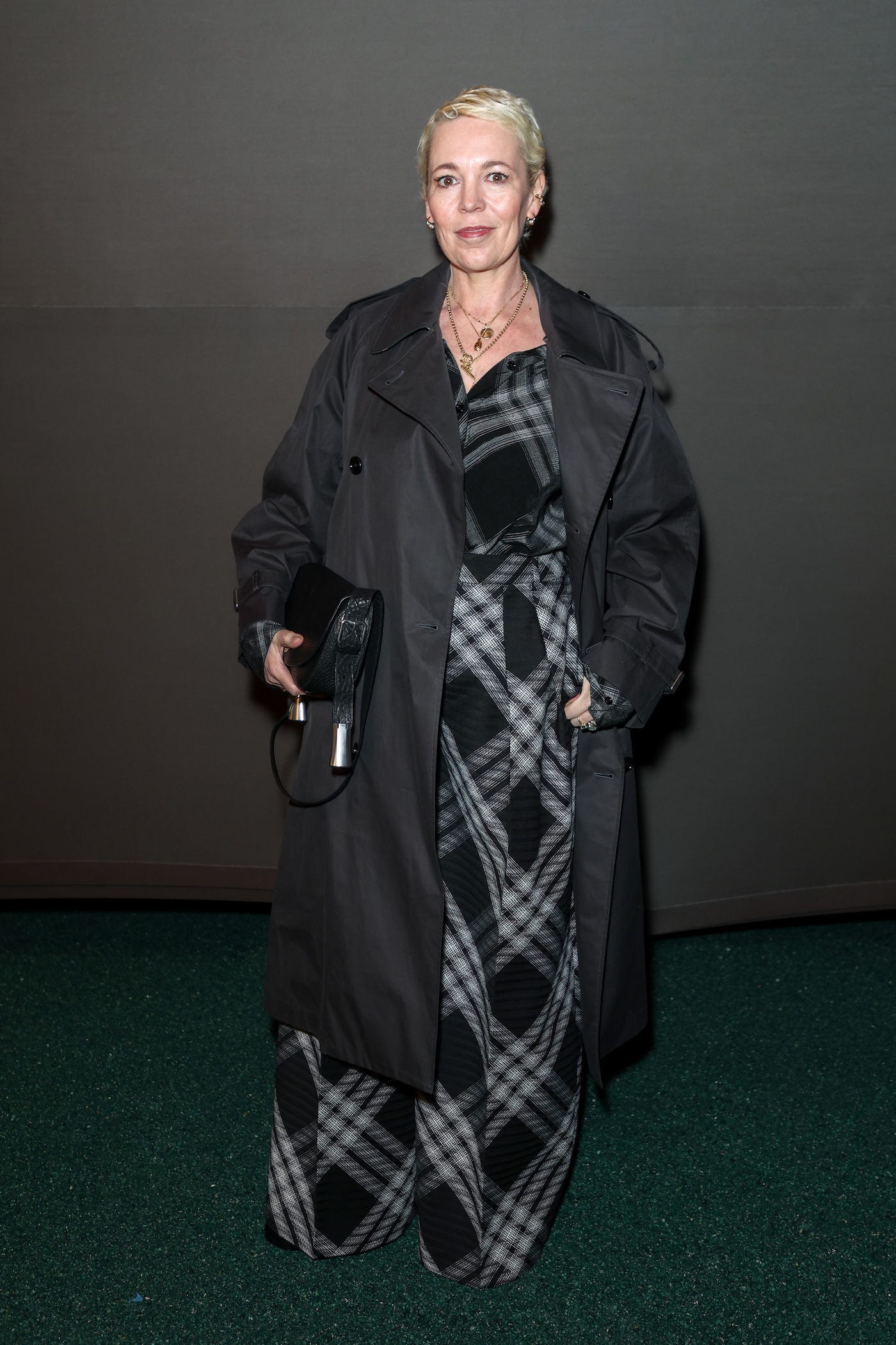 Olivia Colman w eleganckim, czarnym płaszczu oraz wzorzystej sukience w czarną kratę marki Burberry, pozuje na tle ciemnej ściany.