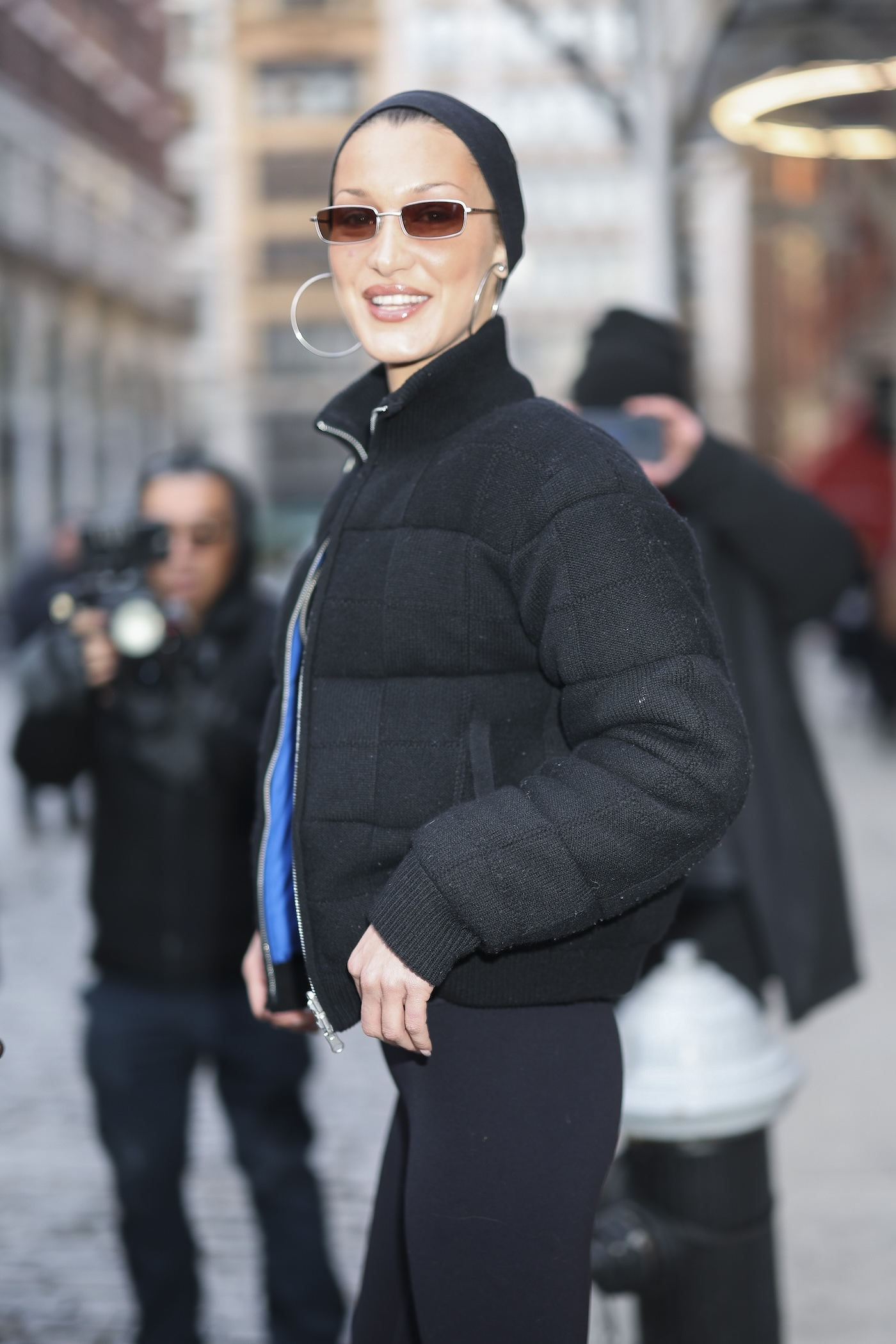 Bella Hadid uśmiecha się do zdjęcia, nosząc czarną kurtkę, opaskę na włosach, duże kolczyki-koła i okulary przeciwsłoneczne podczas spaceru w Nowym Jorku.