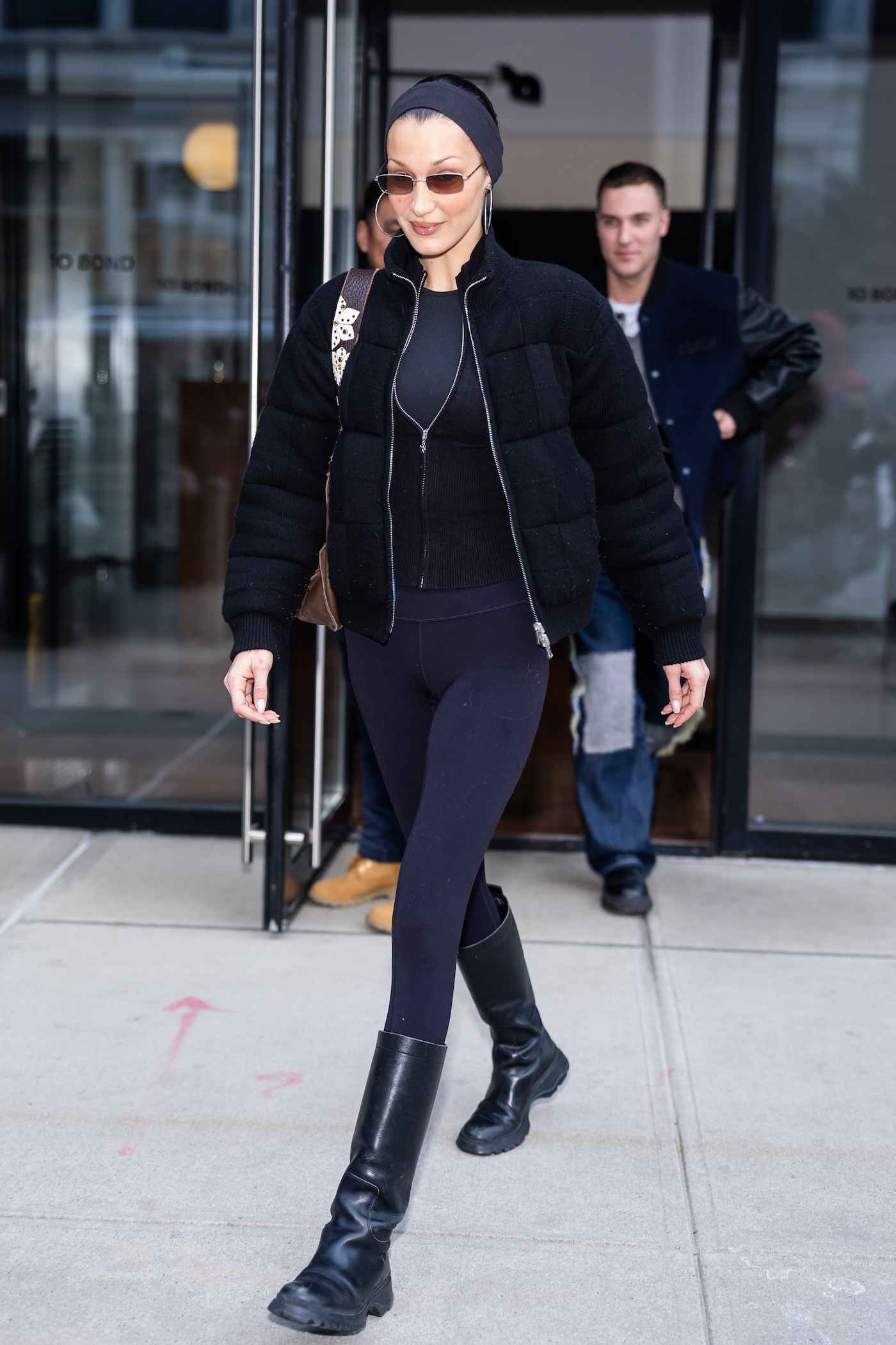 Bella Hadid podczas spaceru w Nowym Jorku nosi legginsy damskie z oficerkami i elegancką wełnianą kurtką, do tego ma czarne, obcisłe spodnie, okulary przeciwsłoneczne oraz opaskę na włosach.