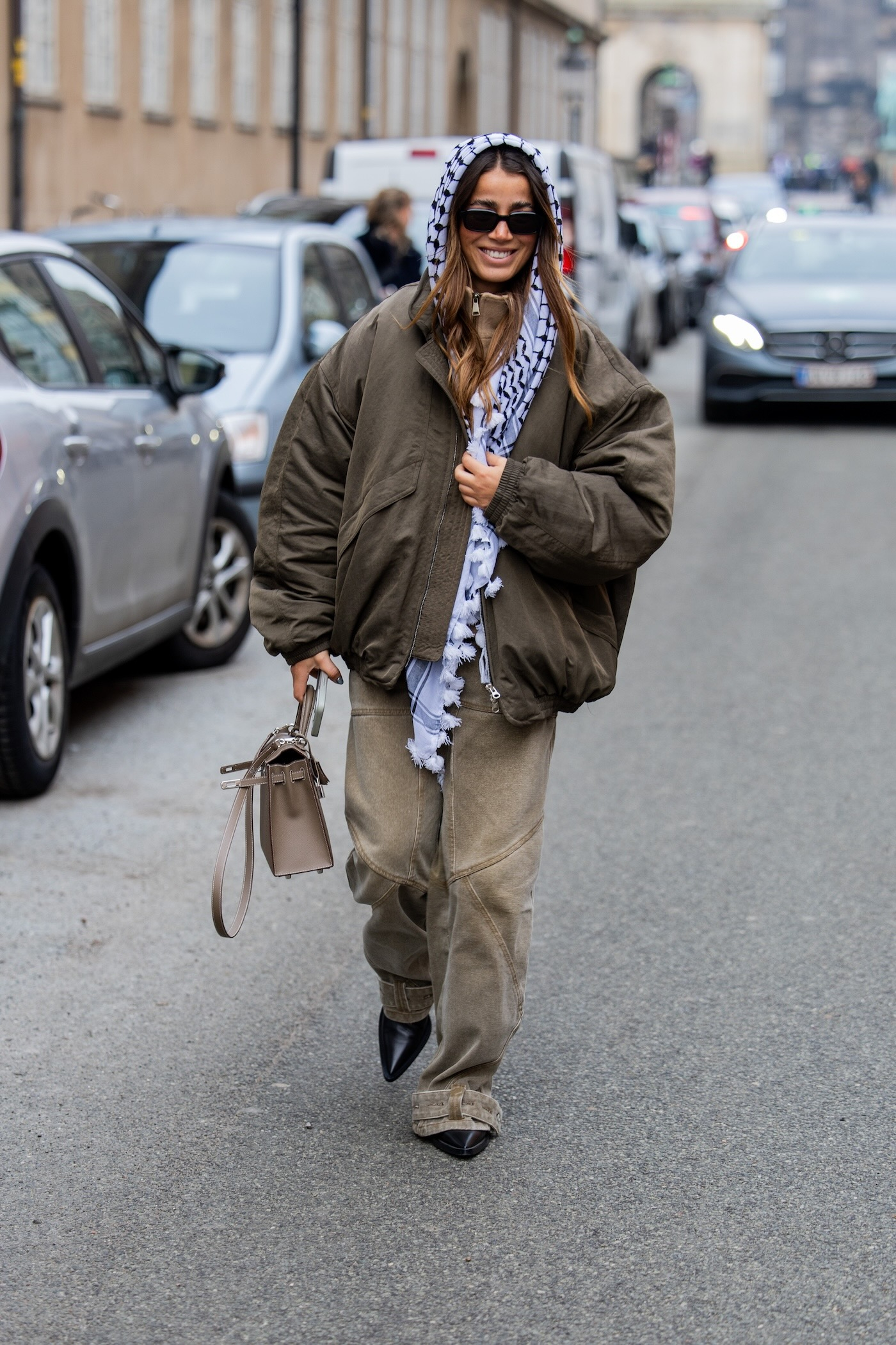 Kobieta sfotografowana podczas Copenhagen Fashion Week w Kopenhadze, ma na sobie szalik, kurtkę bomberkę, bladozielone spodnie i torbę Hermes.