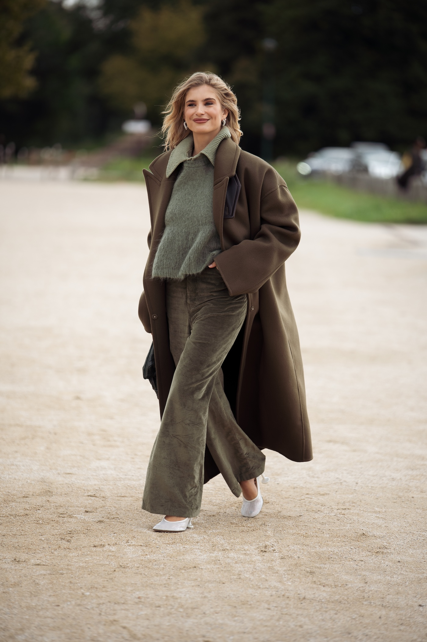 Xenia Adonts nosi oliwkowozielone spodnie, zielony sweter, ciemnozielony płaszcz oversize i białe buty na obcasie podczas Paris Fashion Week w Paryżu.