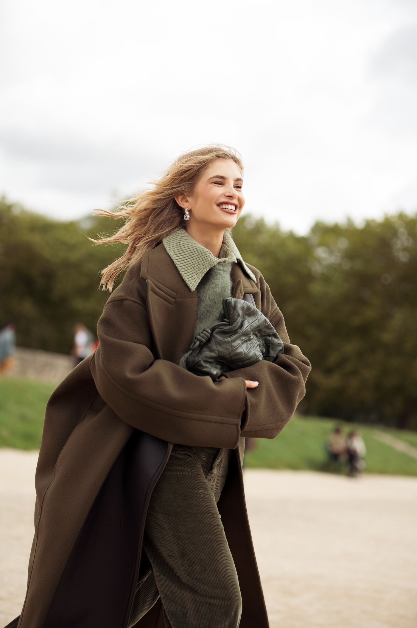 Xenia Adonts nosi oliwkowozielone spodnie, zielony sweter, ciemnozielony płaszcz oversize i białe buty na obcasie podczas Paris Fashion Week w Paryżu.