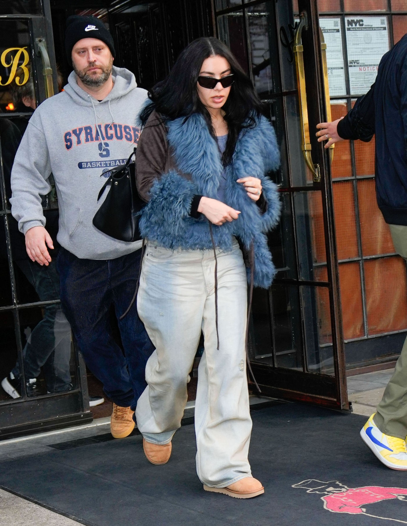 Charli XCX w brązowych butach Ugg, jasnych baggy jeans i awangardowej kurtce z niebieskim futerkiem.