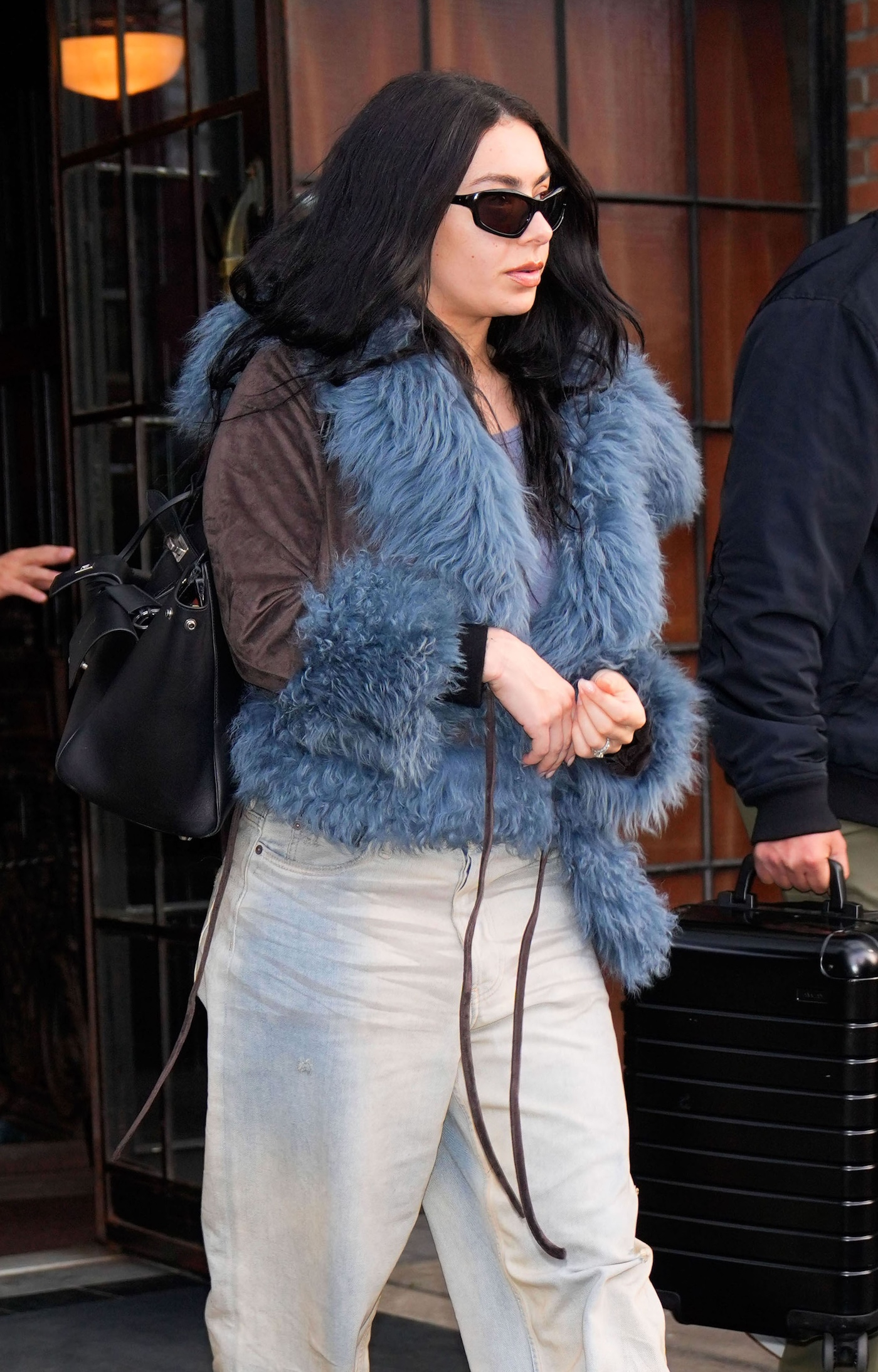 Charli XCX w czarnych okularach przeciwsłonecznych,  jasnych baggy jeans i kurtce z niebieskim futerkiem.