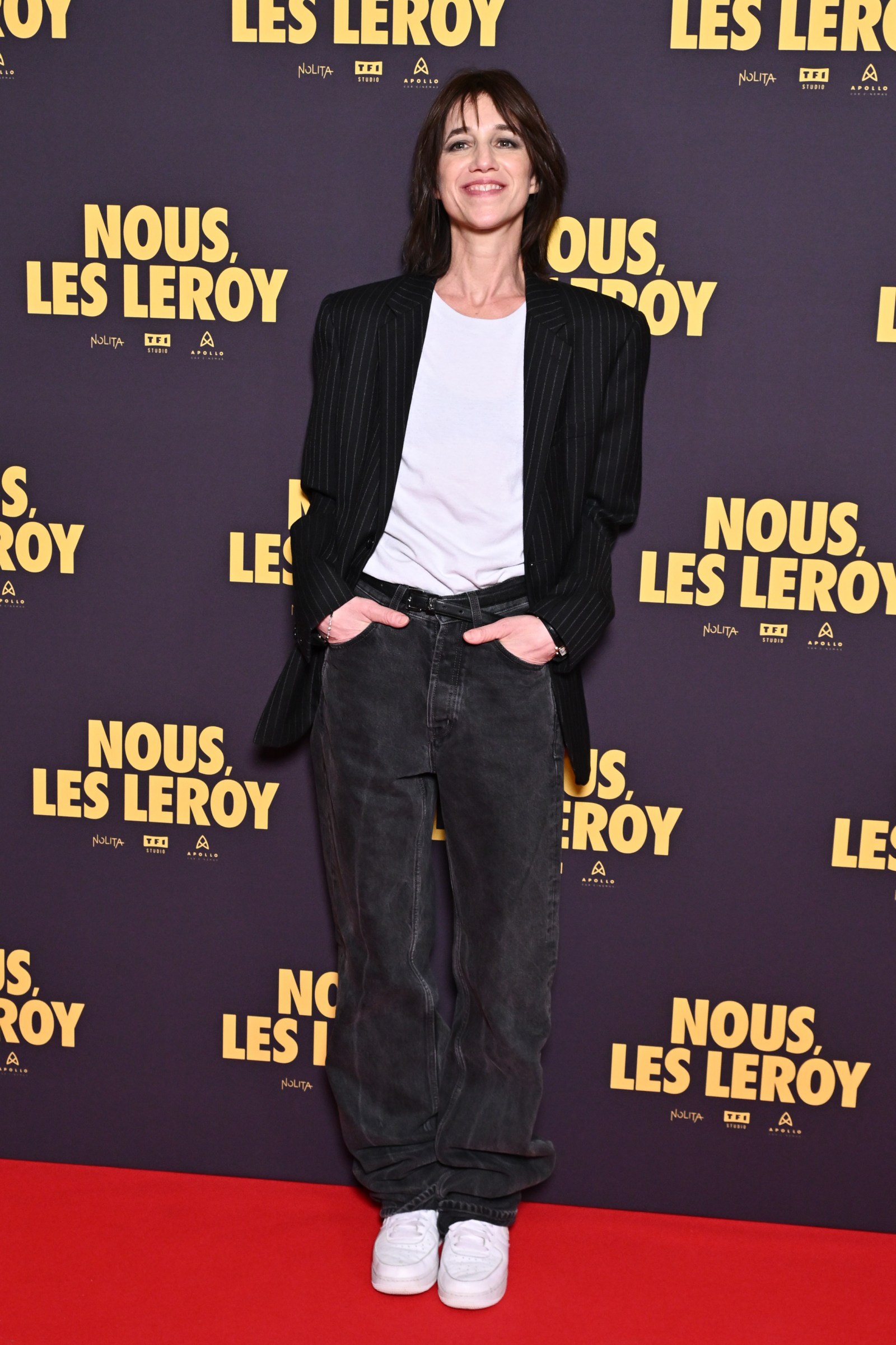 Charlotte Gainsbourg w baggy jeans i sneakersach, stoi na tle plakatu filmowego "Nous, les Leroy".