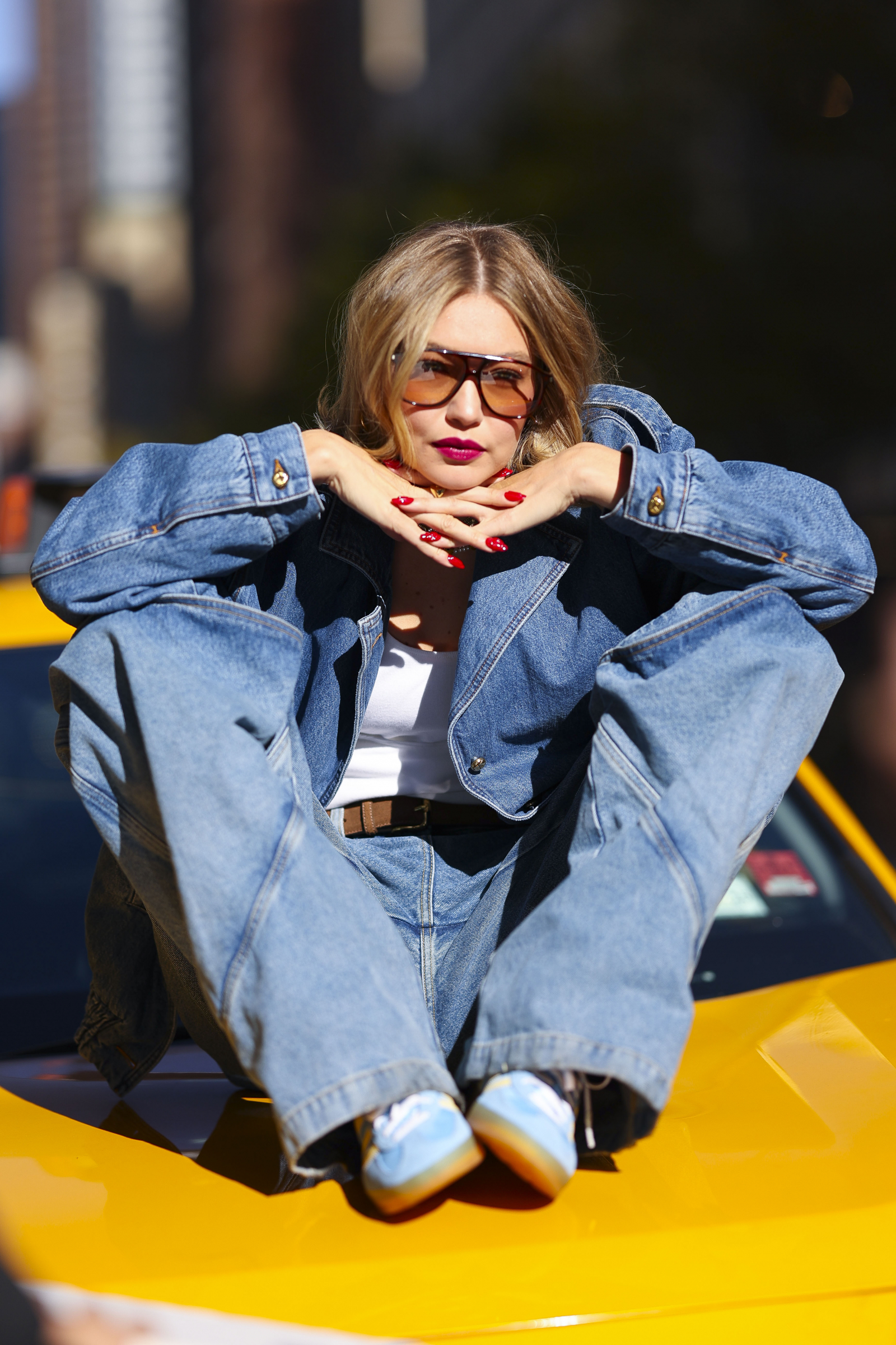 Gigi Hadid w baggy jeans i sneakersach, siedząca na dachu żółtej taksówki, w okularach przeciwsłonecznych.