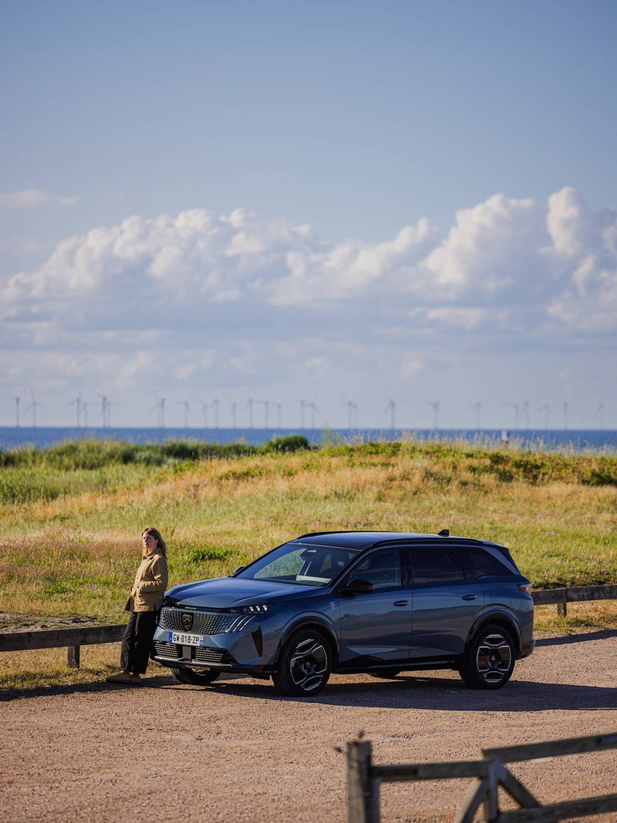 Kobieta stojąca na tle łąki i morza, oparta o samochód Peugeot E-5008