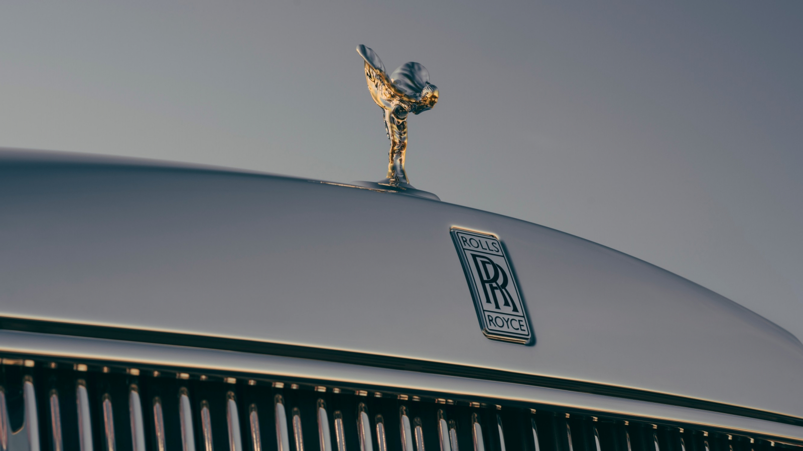 Maskownica samochodu Rolls-Royce z figurką Spirit of Ecstasy oraz logo marki na tle nieba.