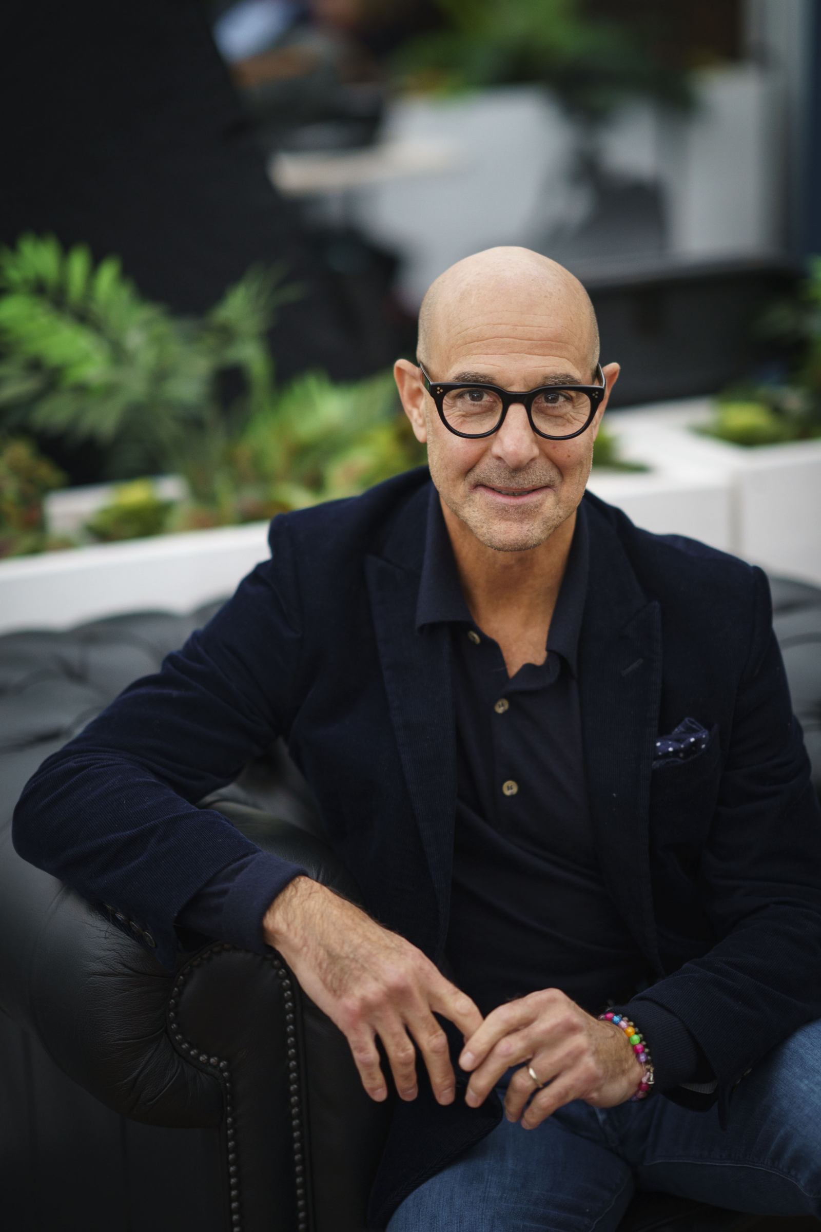 Stanley Tucci siedzi na ciemnej kanapie, uśmiechnięty, z okularami i w eleganckim ubraniu, otoczony roślinami.