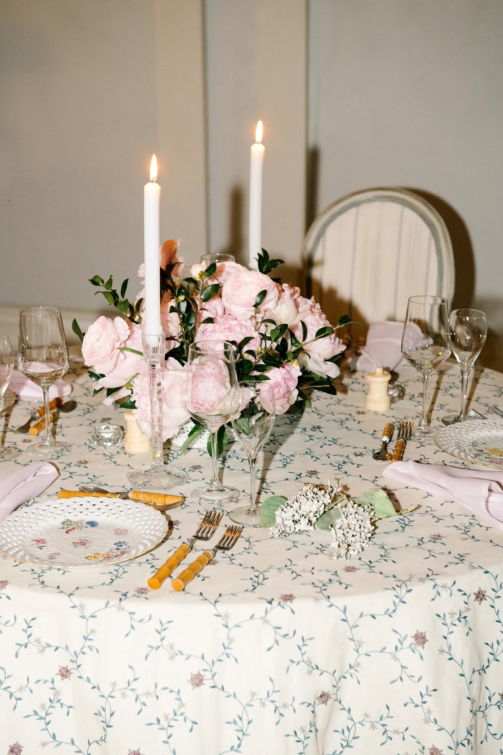 Stół z kwiatową dekoracją, świecami i eleganckimi naczyniami na obrusie w kwiatowy wzór.