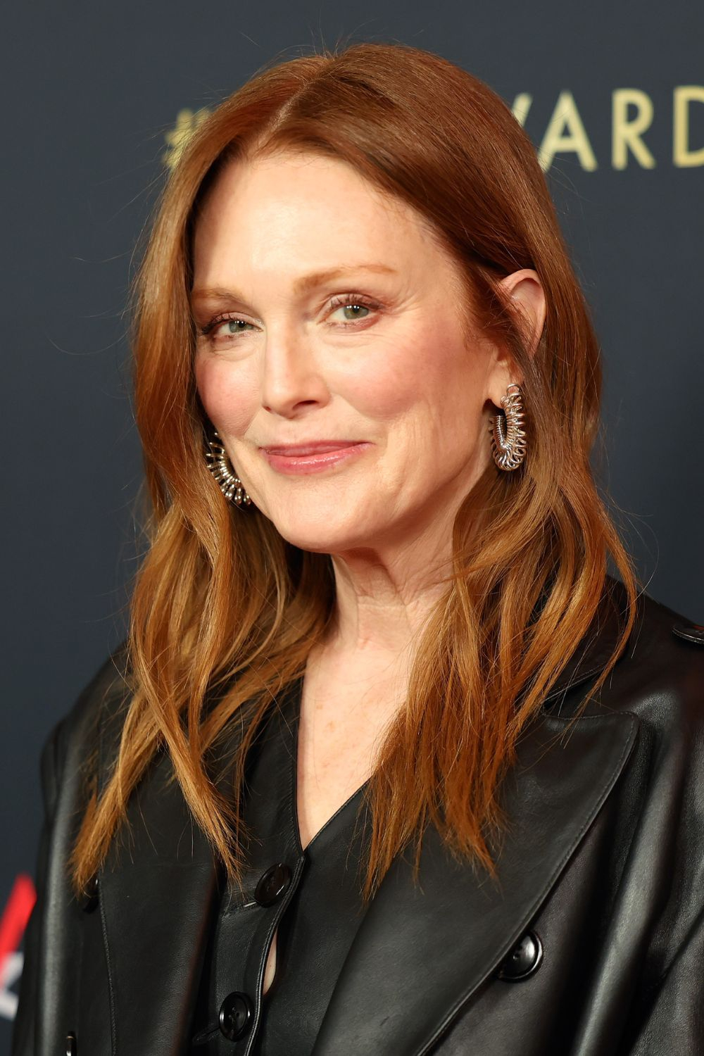 Julianne Moore w masywnych i długich kolczykach, w czarnej kurtce, uśmiechnięta na tle z napisem.