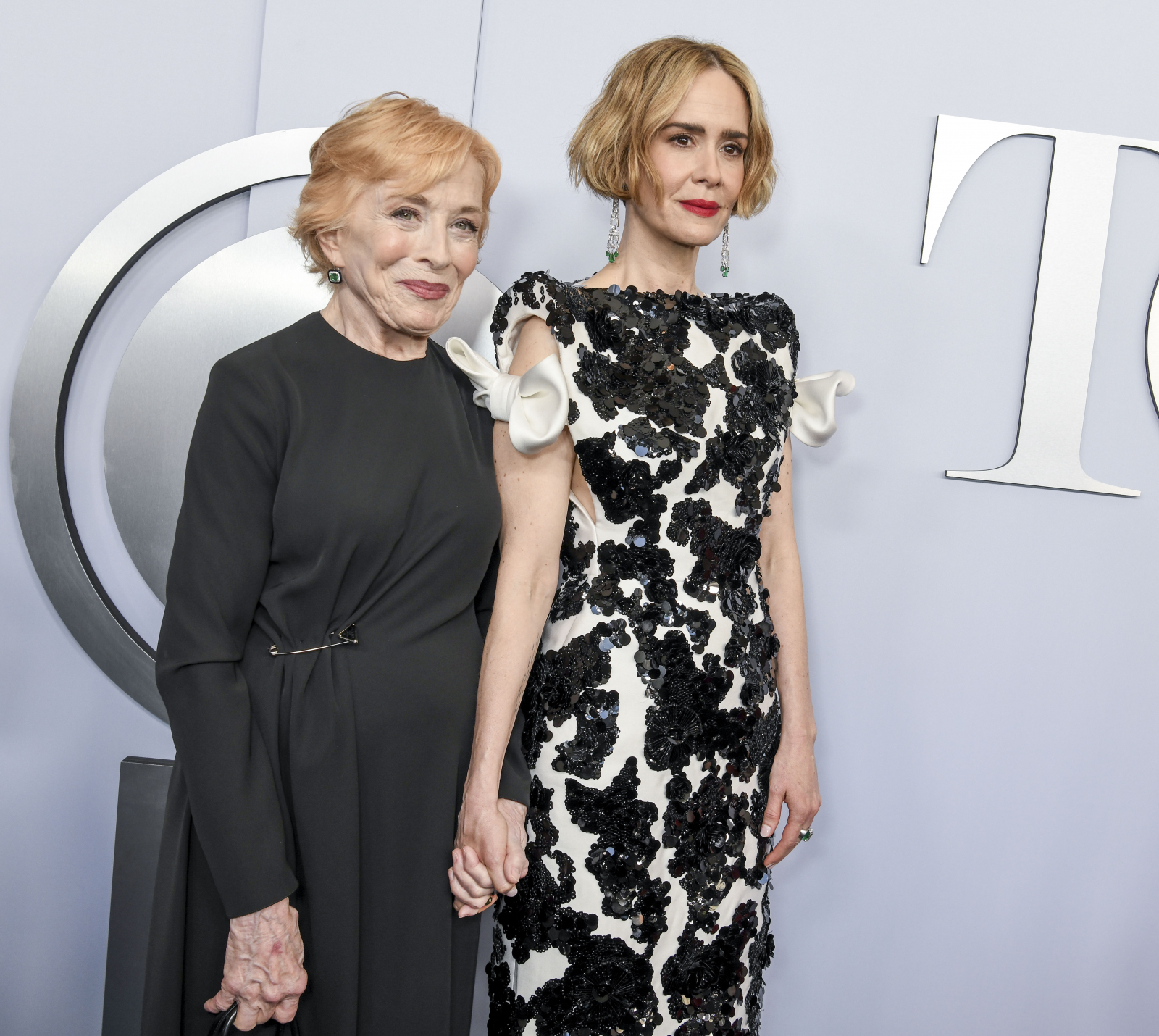 Sarah Paulson i Holland Taylor trzymają się za ręce, pozując na tle eleganckiego tła. Obie ubrane w stylowe kreacje.