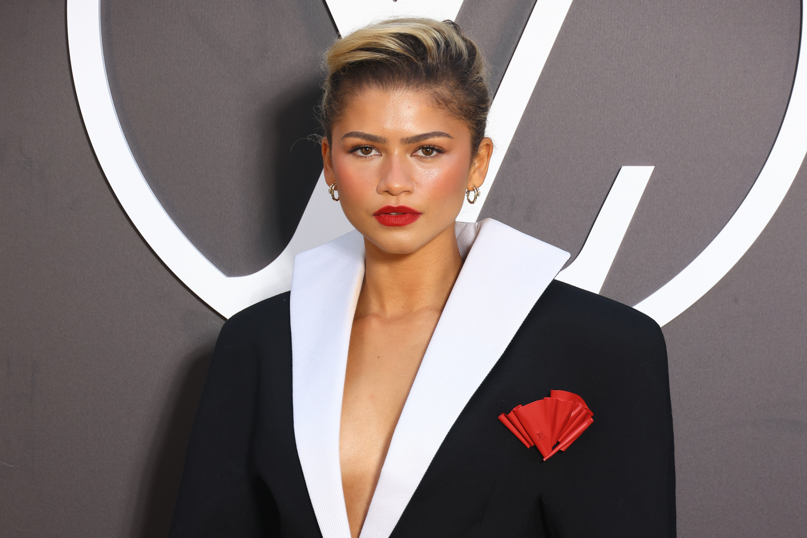 Zendaya w eleganckim czarnym garniturze z białymi klapami i czerwonym akcentem, z wyrazistym makijażem.